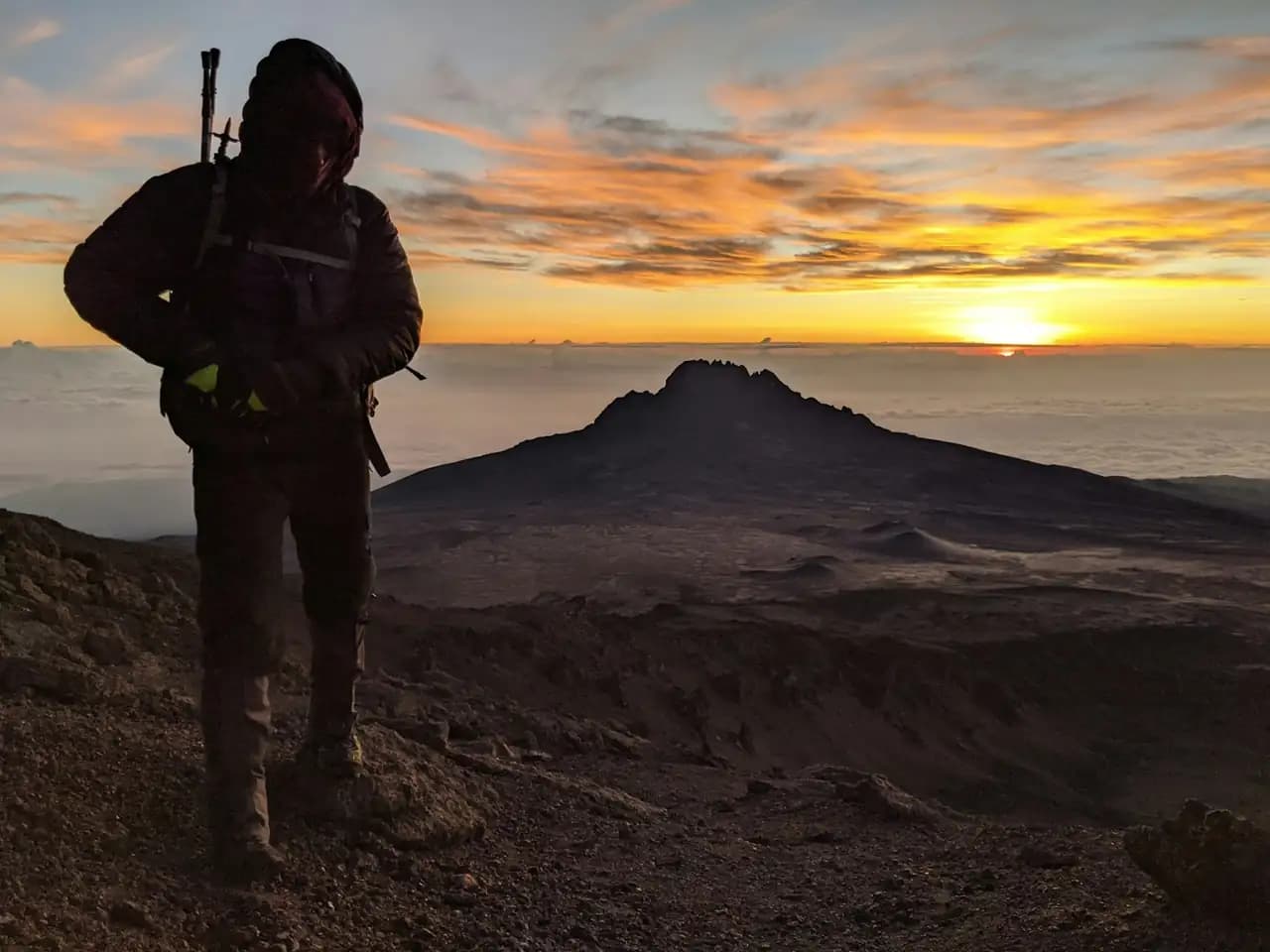 A trekker standing at sunrise above the clouds on Kilimanjaro, with the jagged Mawenzi peak silhouetted against an orange sky